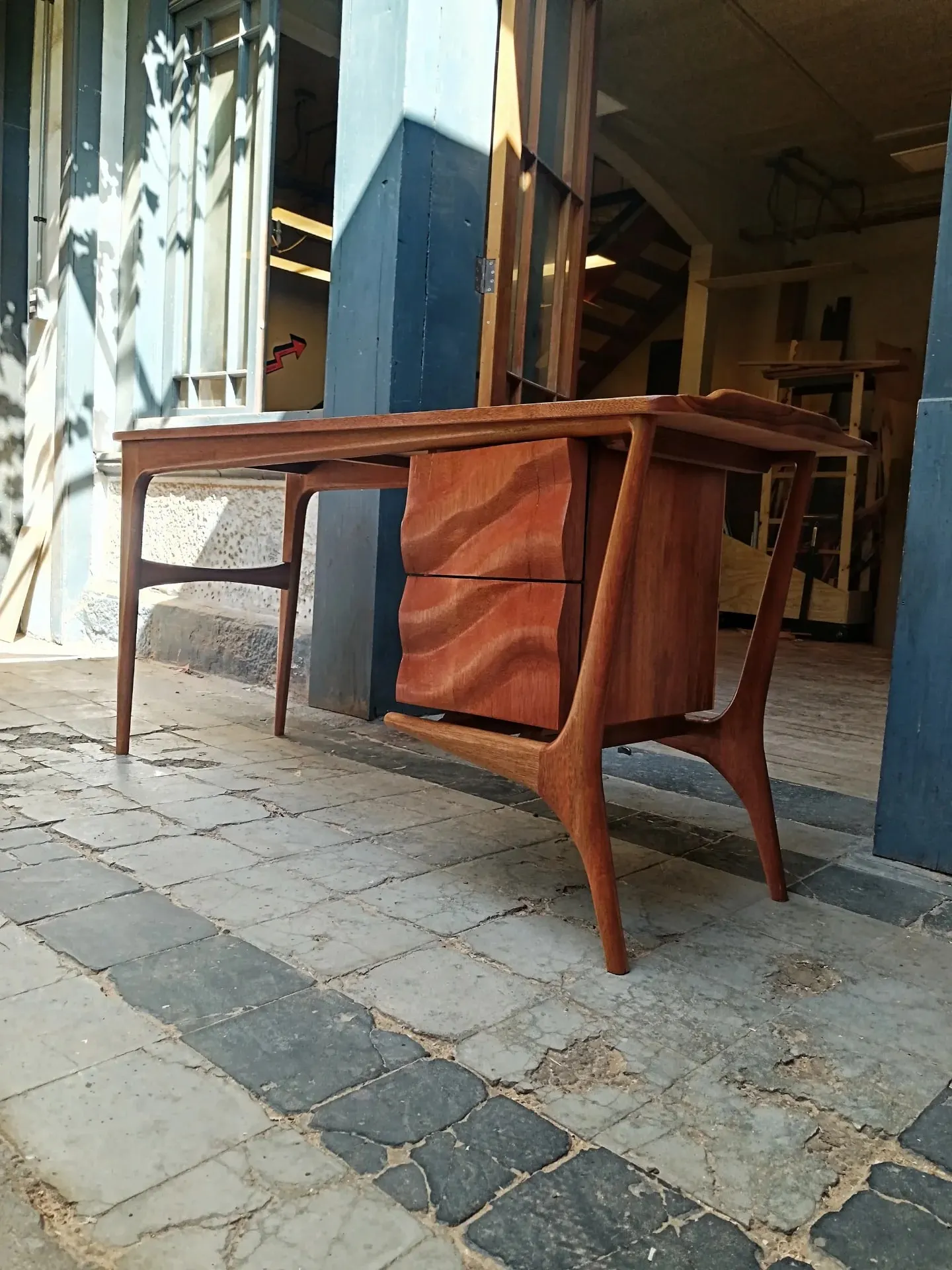 Escritorio de madera rojiza de estilo mid-century, con dos cajones tallados en ondas y patas esculpidas, a la luz del sol en la entrada del taller.