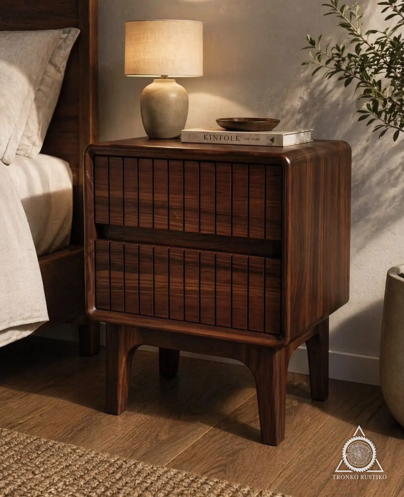 Velador de madera oscura de cantos curvos con dos cajones de frente ranurado en vertical, sobre patas, junto a una cama.