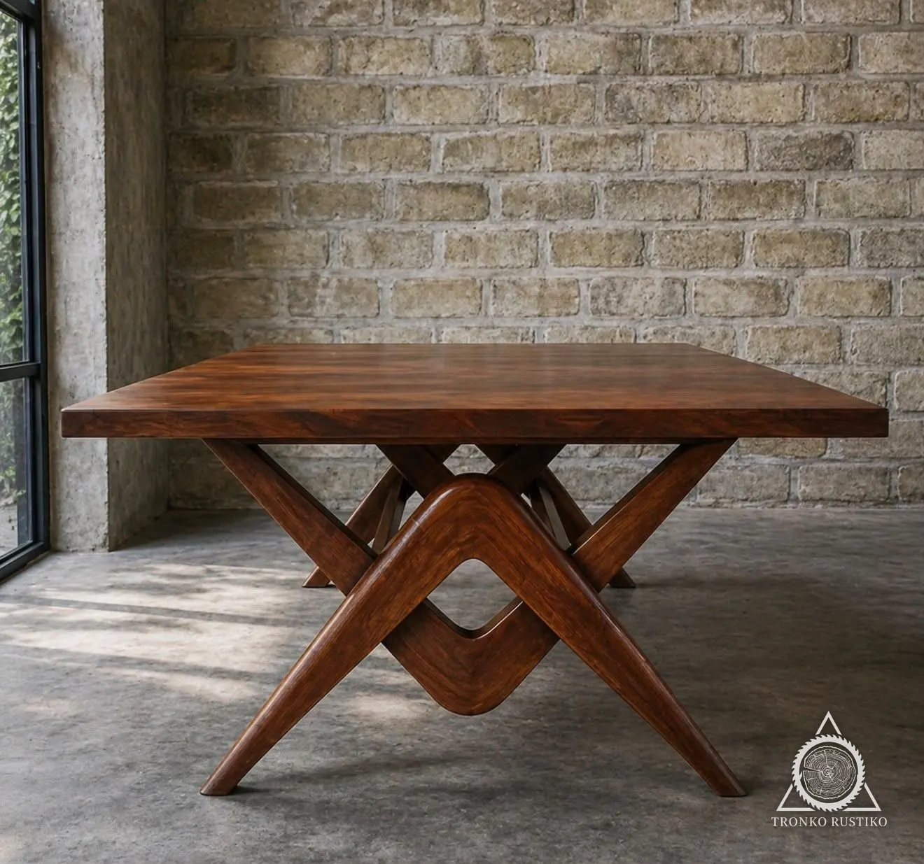 Mesa de comedor de madera rojiza con una base escultórica de piezas curvas entrecruzadas, frente a un muro de ladrillo.