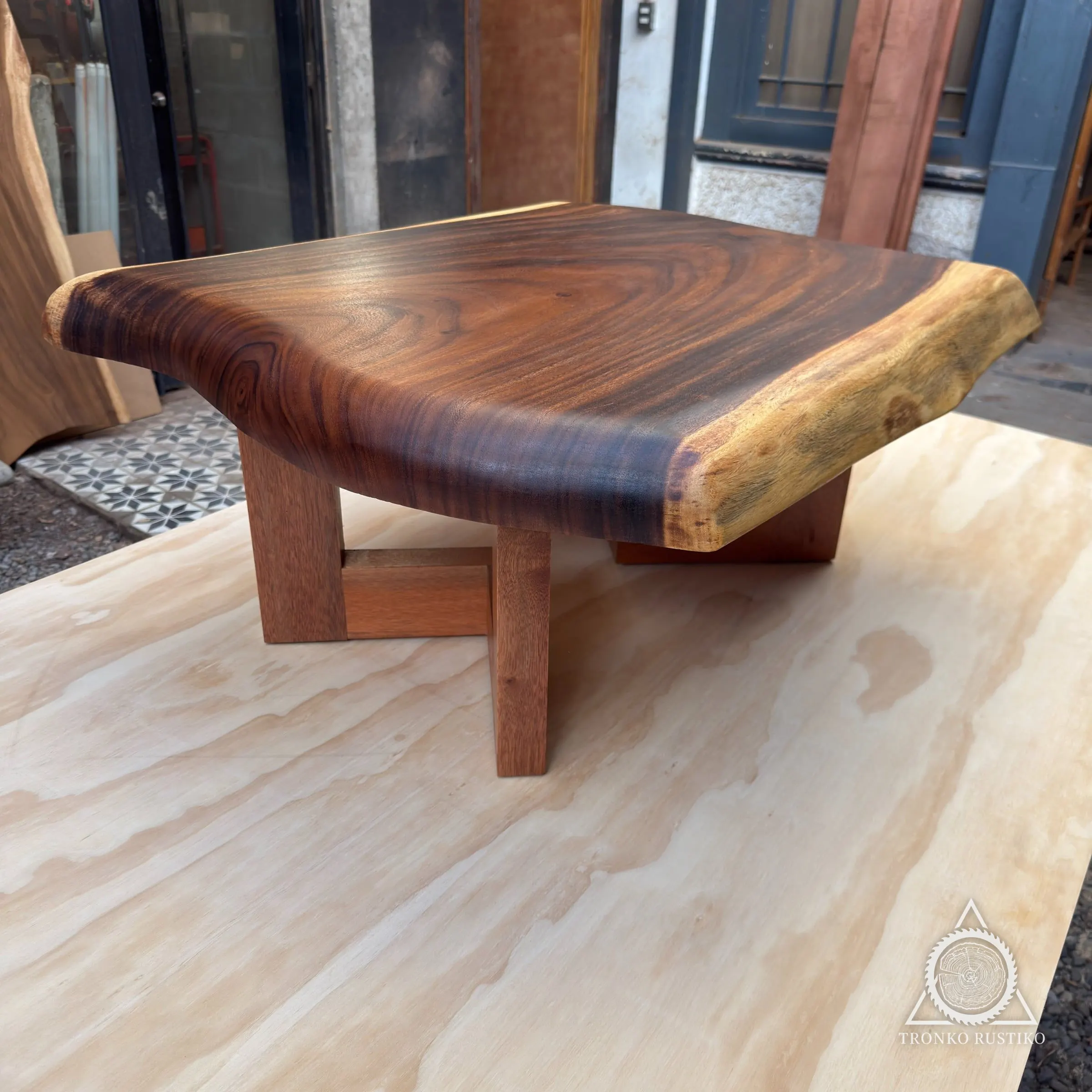 Mesa de centro con cubierta gruesa de madera oscura de borde natural y canto claro, sobre bases macizas, en el patio del taller.