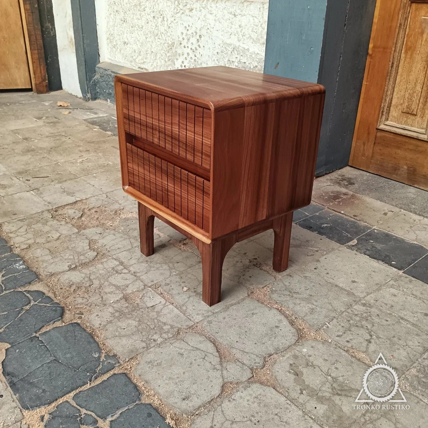 Velador de madera oscura de cantos curvos con dos cajones de frente ranurado y patas, en el patio del taller.