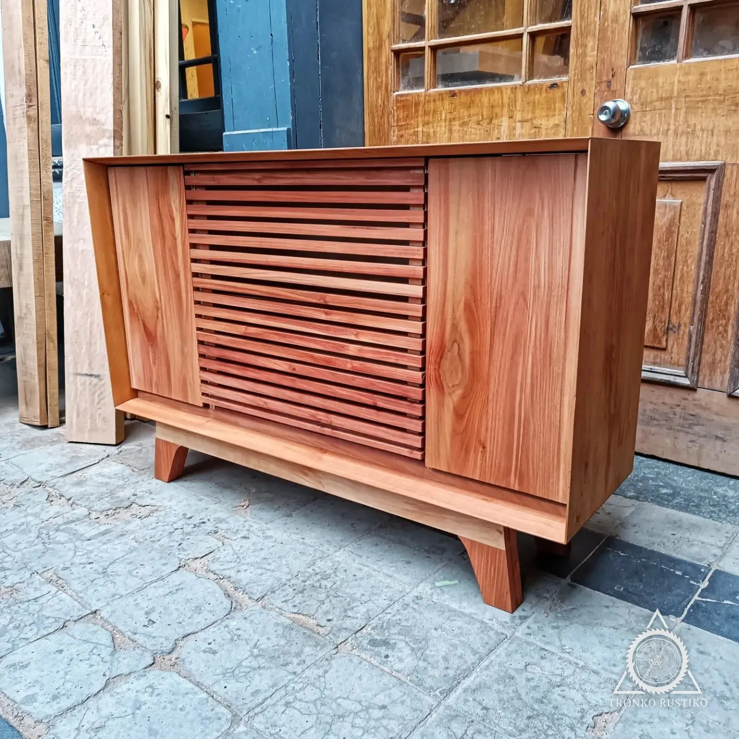 Rack de madera rojiza con palillaje horizontal al centro y patas inclinadas, frente a una puerta del taller.