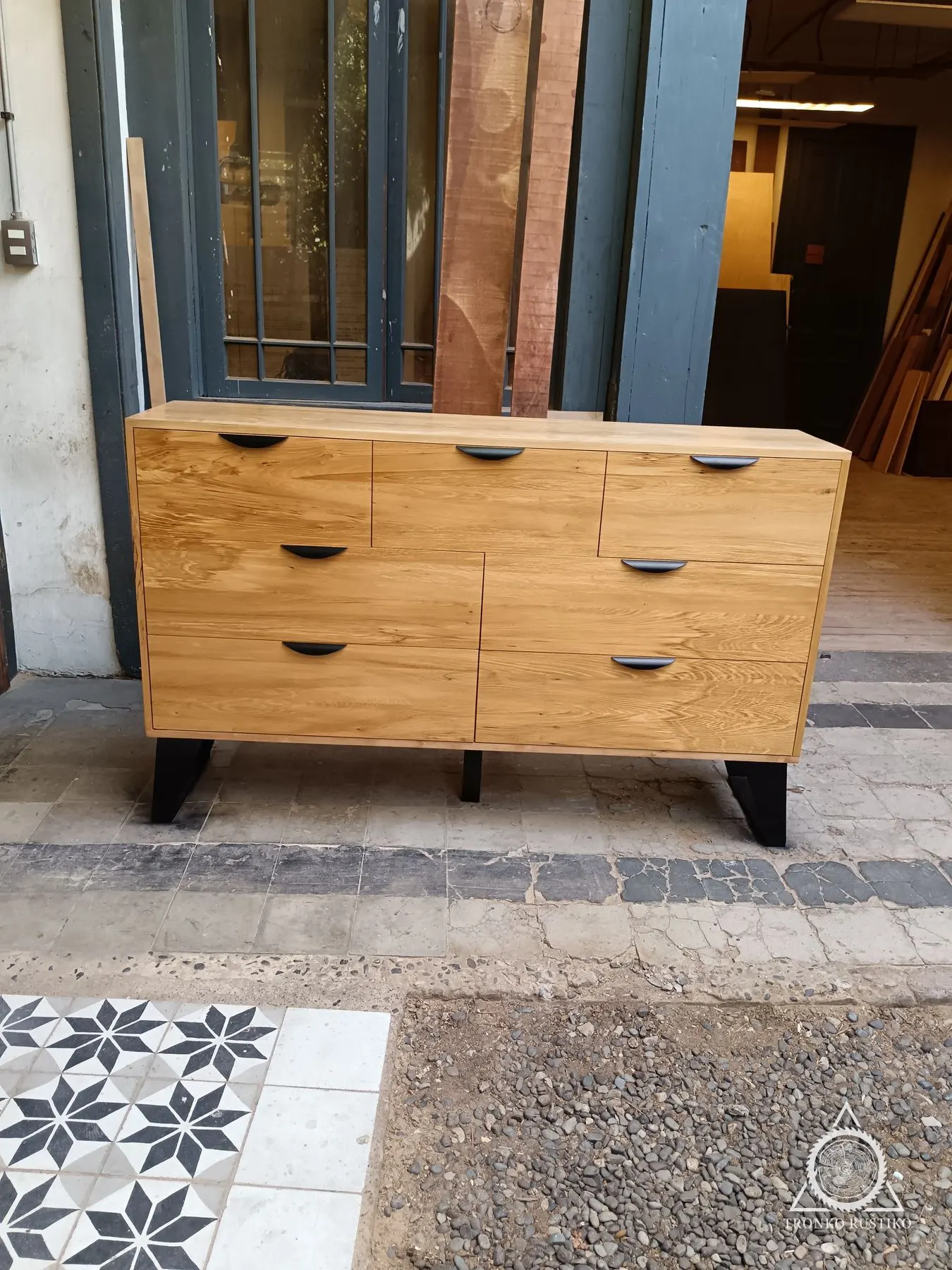 Cómoda de madera clara con cajones de tiradores rebajados y base metálica negra.