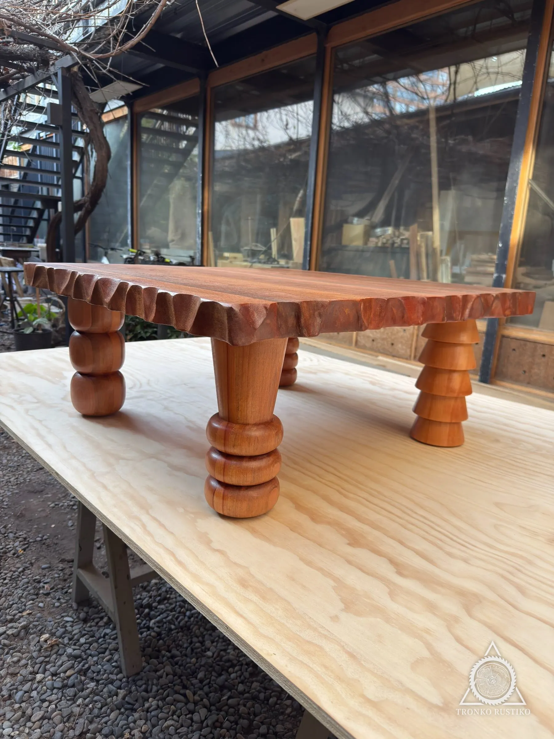 Mesa de centro de madera rojiza con borde tallado ondulado y patas torneadas, sobre un tablero en el patio del taller.