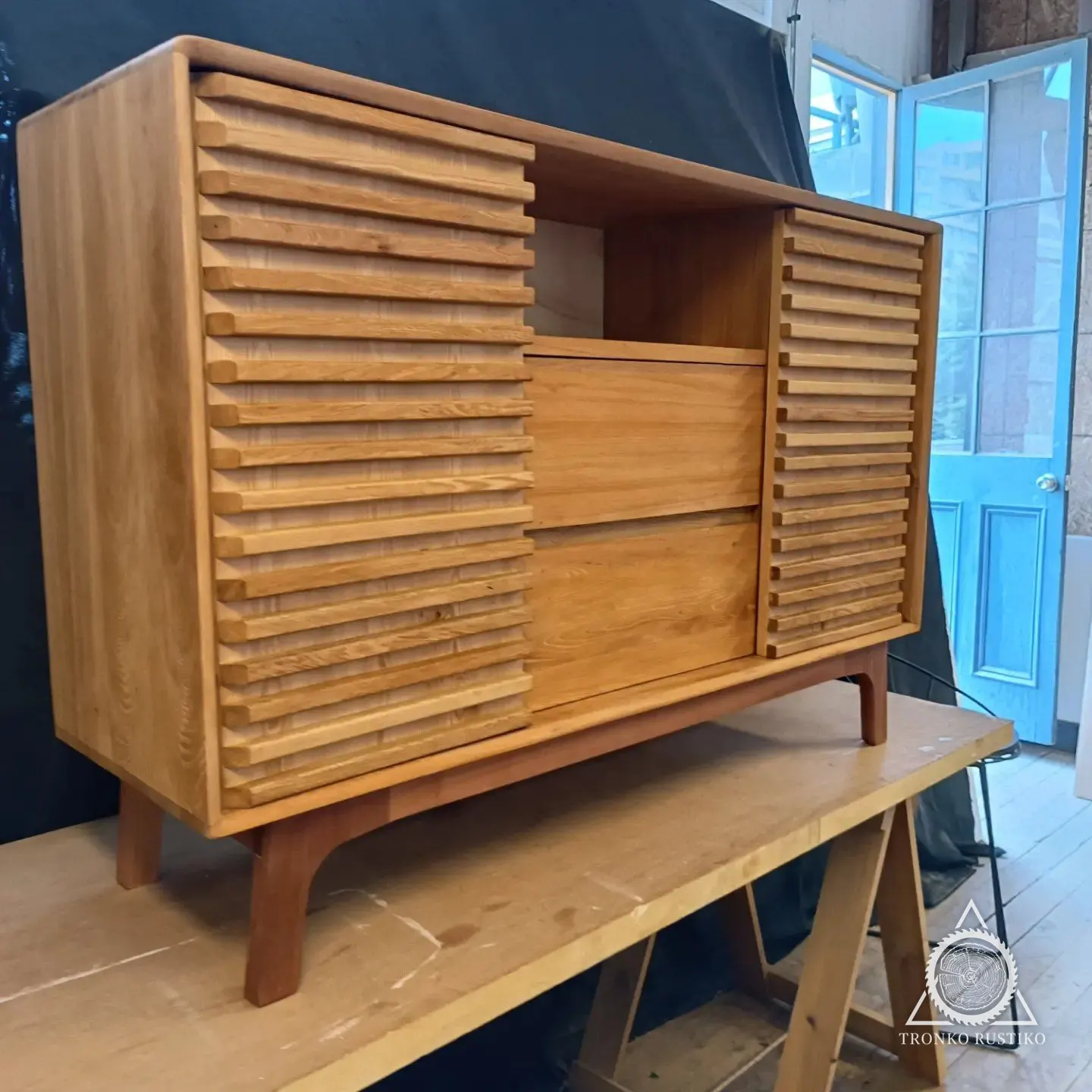 Mueble de madera clara con puertas laterales de listones horizontales, dos cajones al centro y nicho superior, sobre patas inclinadas, en el taller.