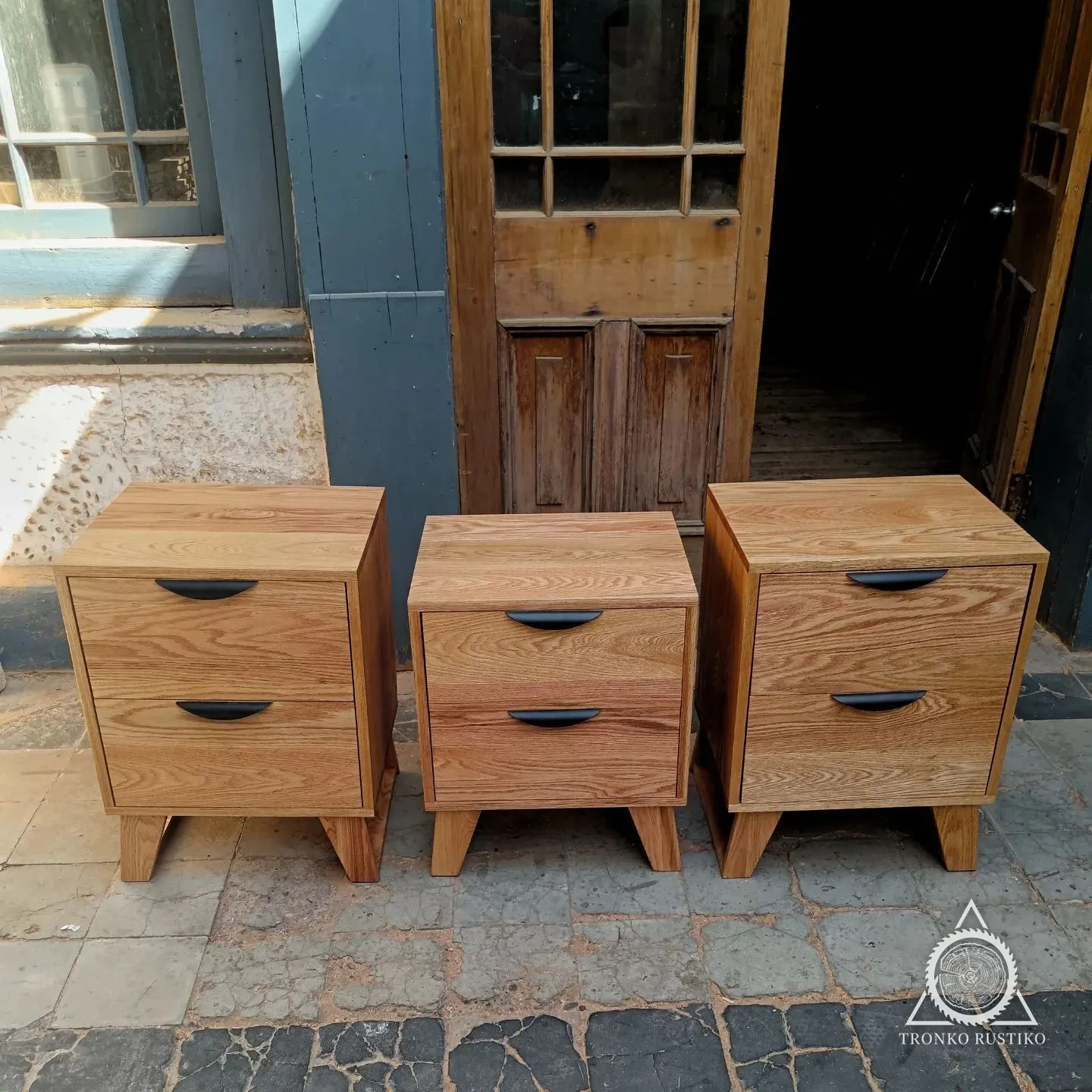 Tres veladores de madera clara con dos cajones y tiradores negros de media luna, sobre patas inclinadas.