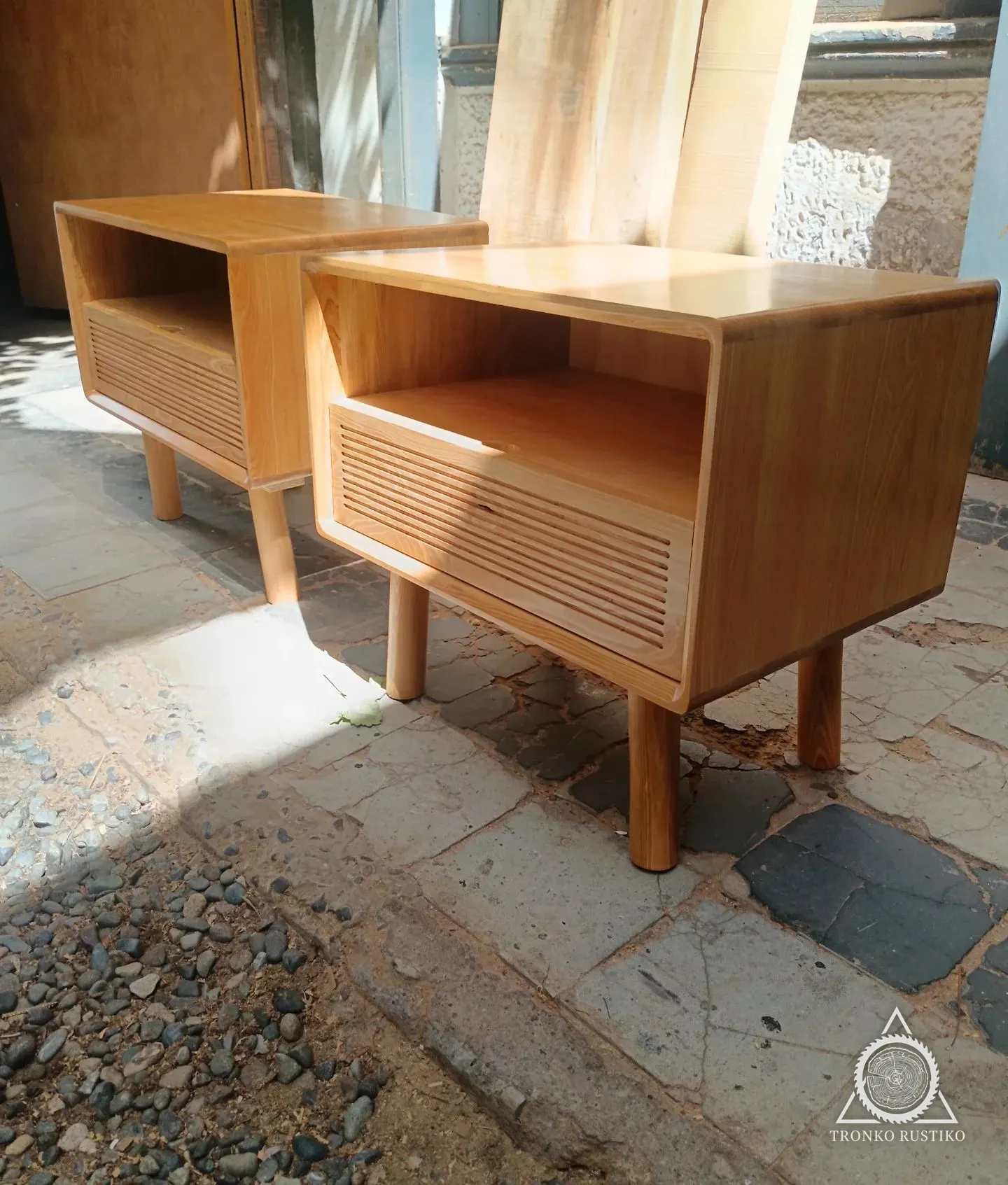 Par de veladores de madera clara con nicho abierto, cajón de frente acanalado y patas cilíndricas.