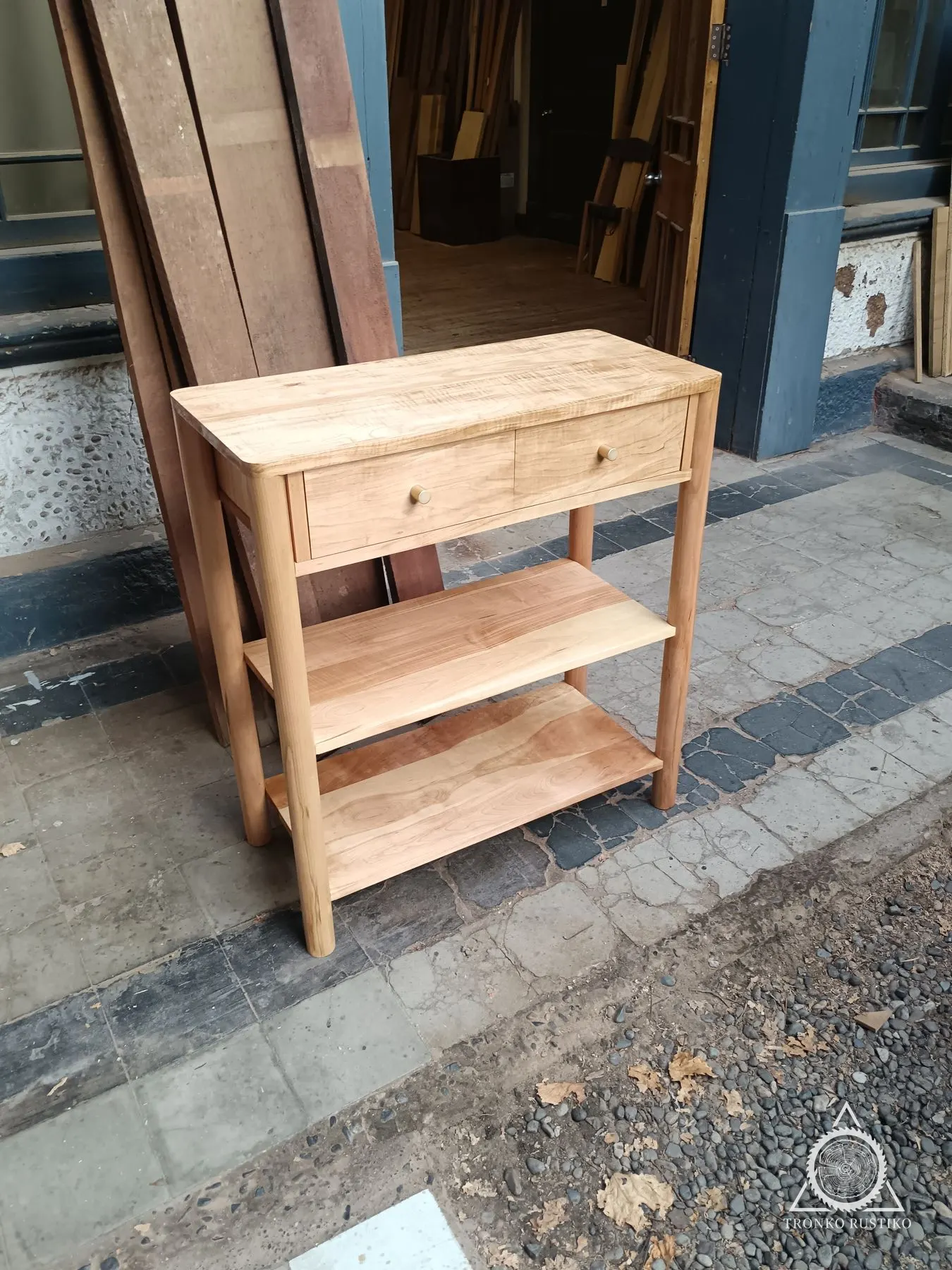 Arrimo de madera clara con cajón superior y dos repisas abiertas, sobre patas rectas.