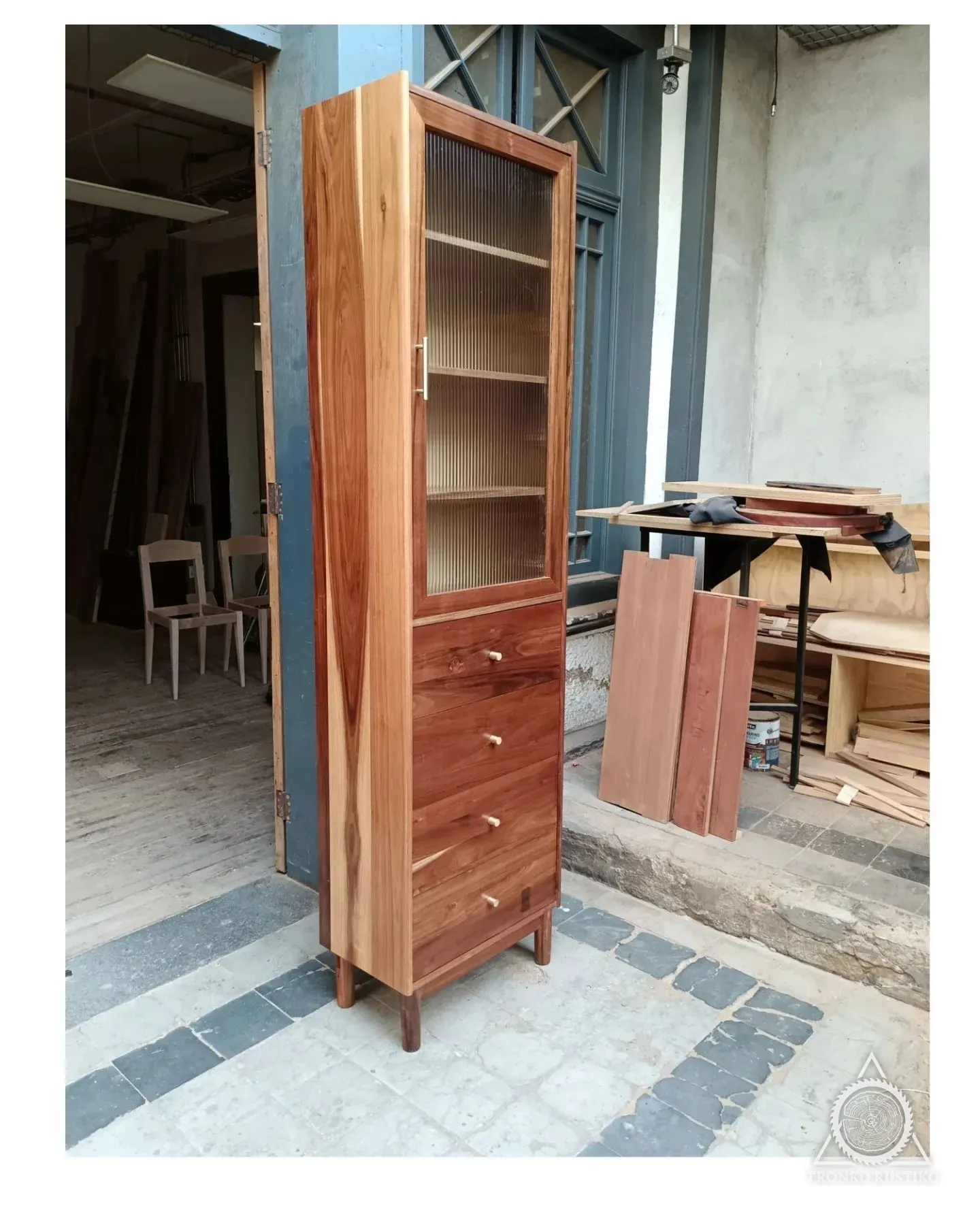 Vitrina alta y angosta de madera con puertas de vidrio acanalado y cajones abajo.