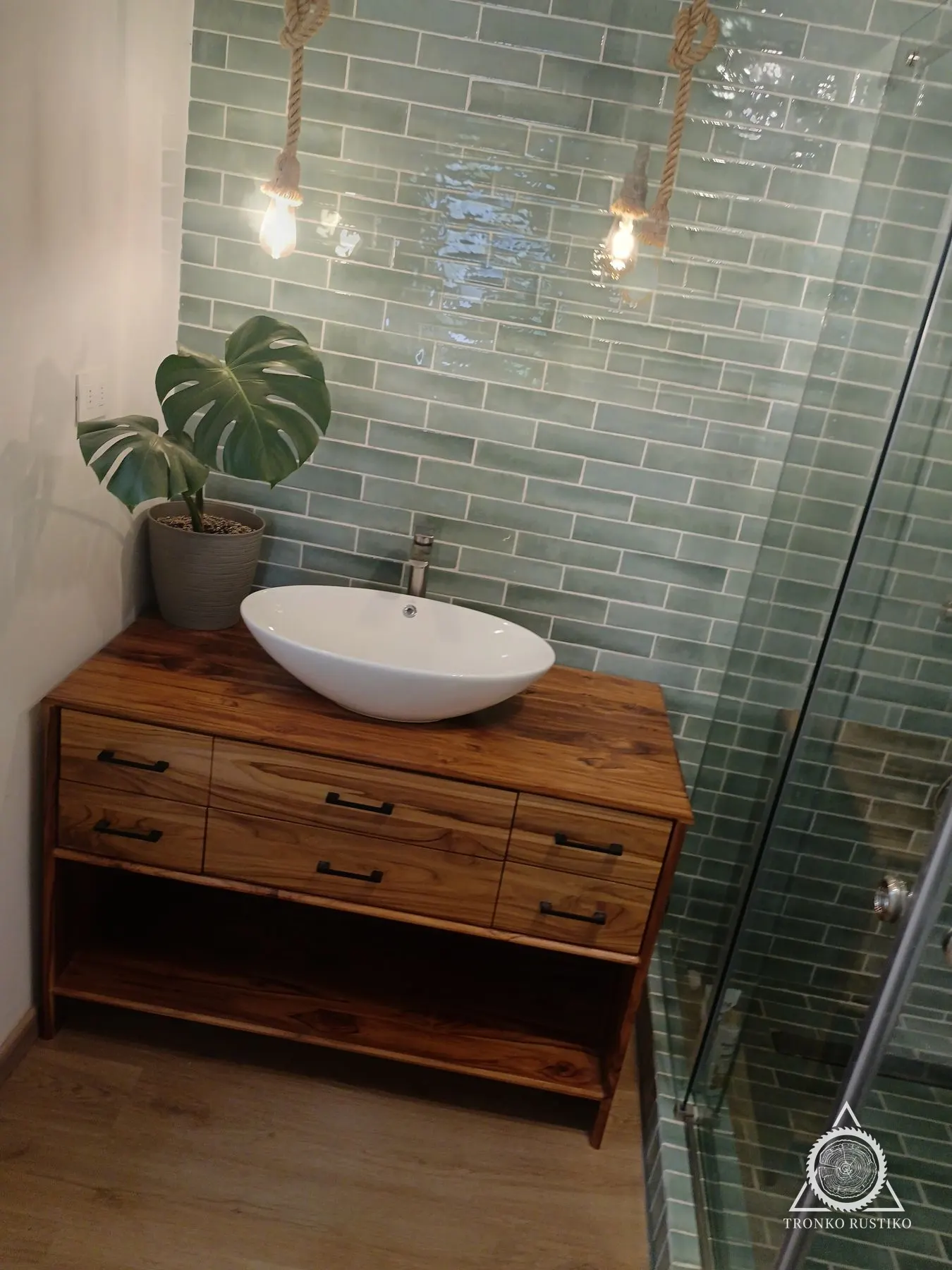 Vanitorio de madera con seis cajones y repisa abierta, con lavamanos blanco sobrepuesto, instalado en un baño de azulejos verdes.