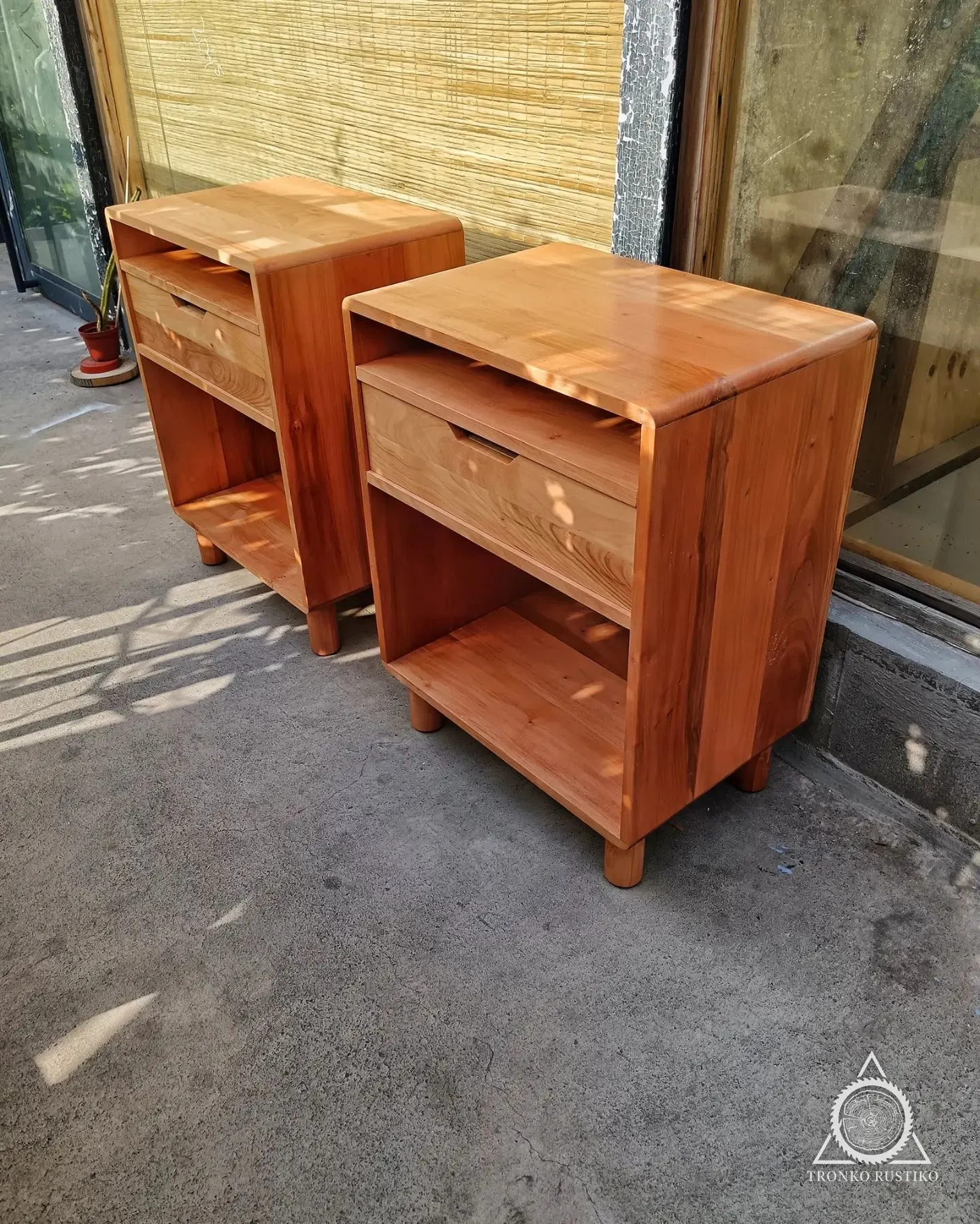 Par de veladores de madera rojiza con un cajón y nichos abiertos, sobre patas redondas.