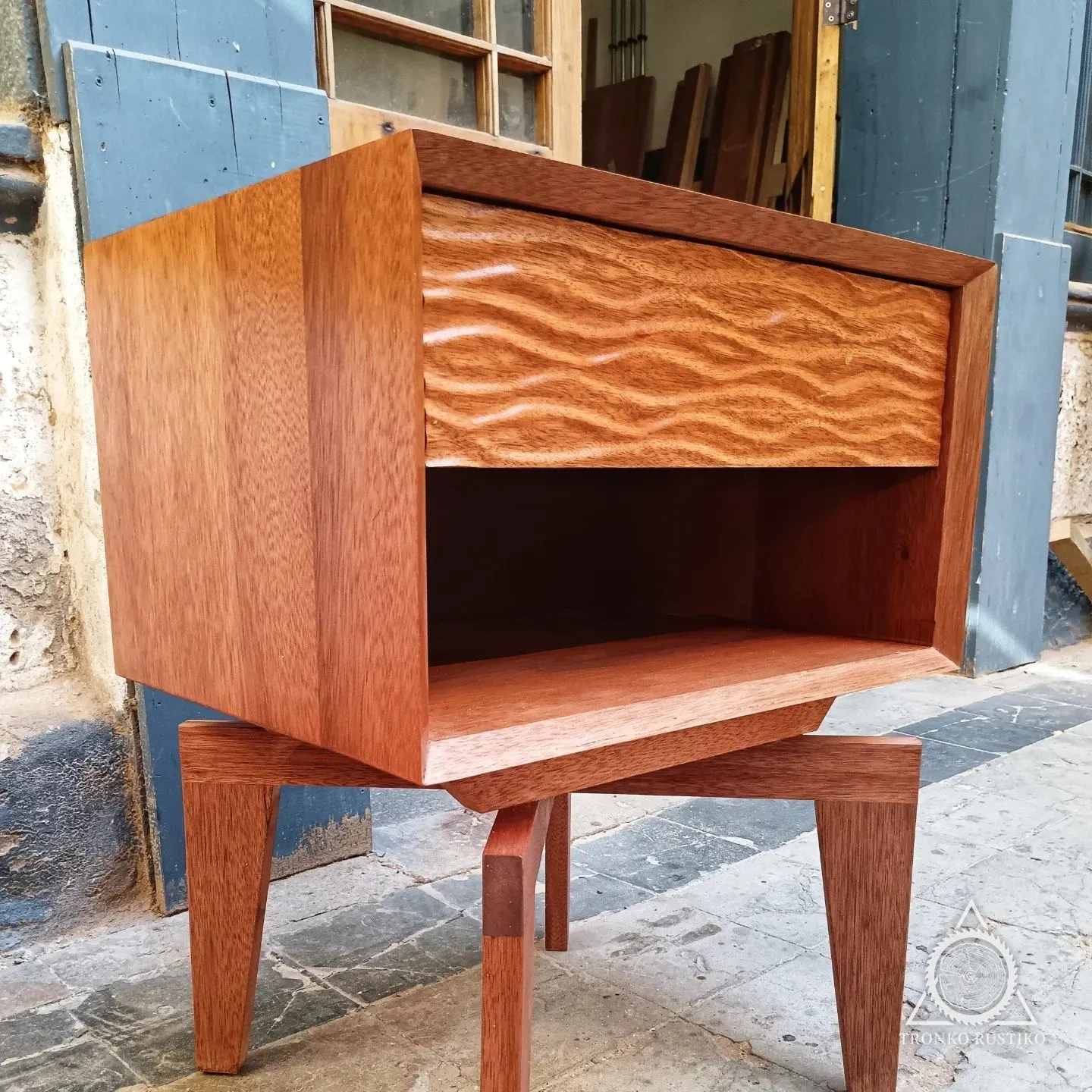 Velador de madera rojiza con cajón de frente tallado en ondas, nicho abierto y base de patas macizas, en el taller.