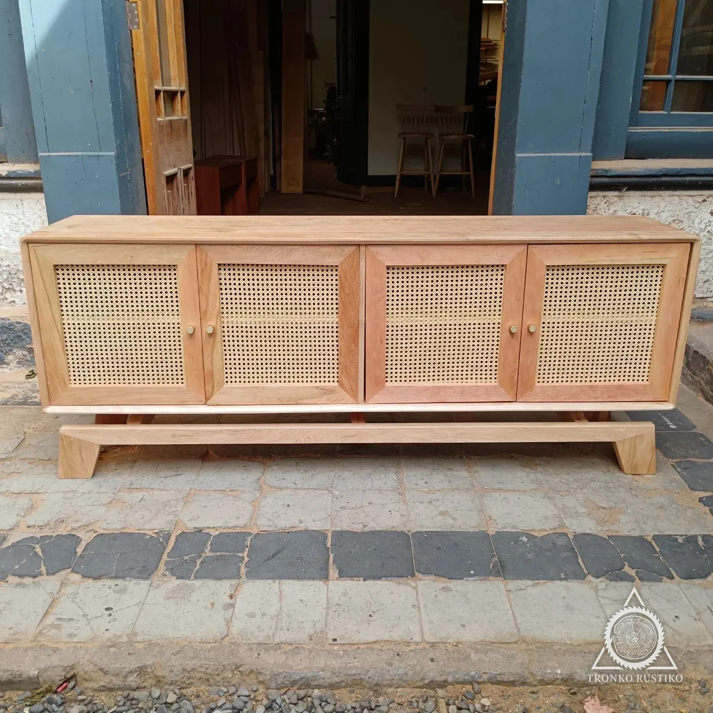 Rack largo de madera clara con cuatro puertas de junco natural y base flotante, en el patio del taller.