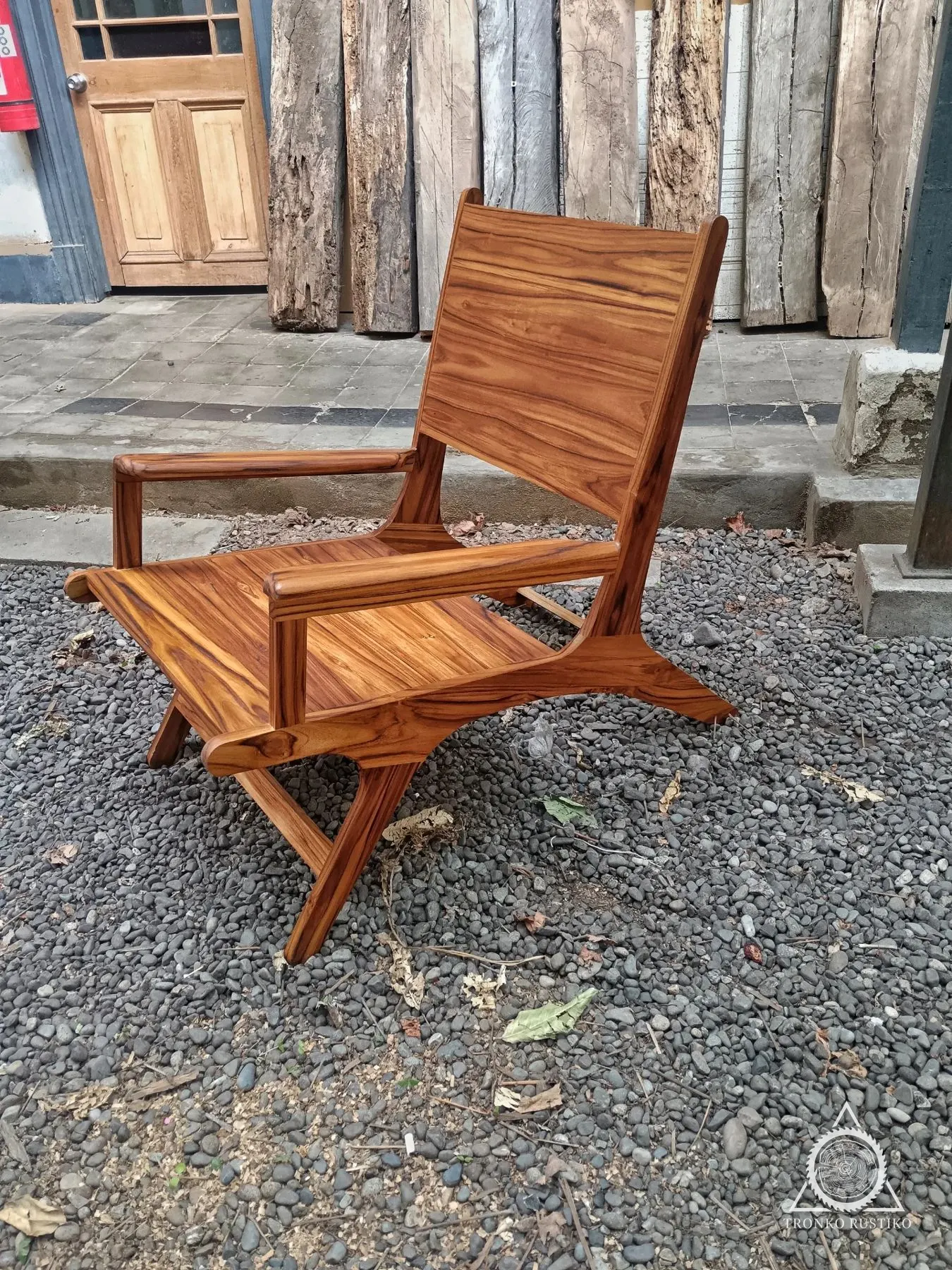 Sillón de descanso de madera con respaldo reclinado y apoyabrazos integrados, sobre un patio de piedras.