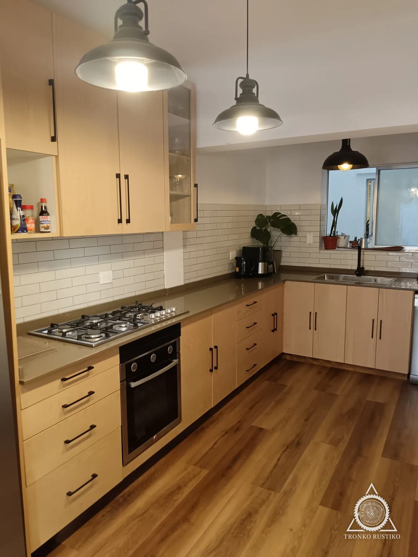 Cocina en L con muebles bajos y altos de frentes color madera rosada, tiradores negros y columna junto al refrigerador.
