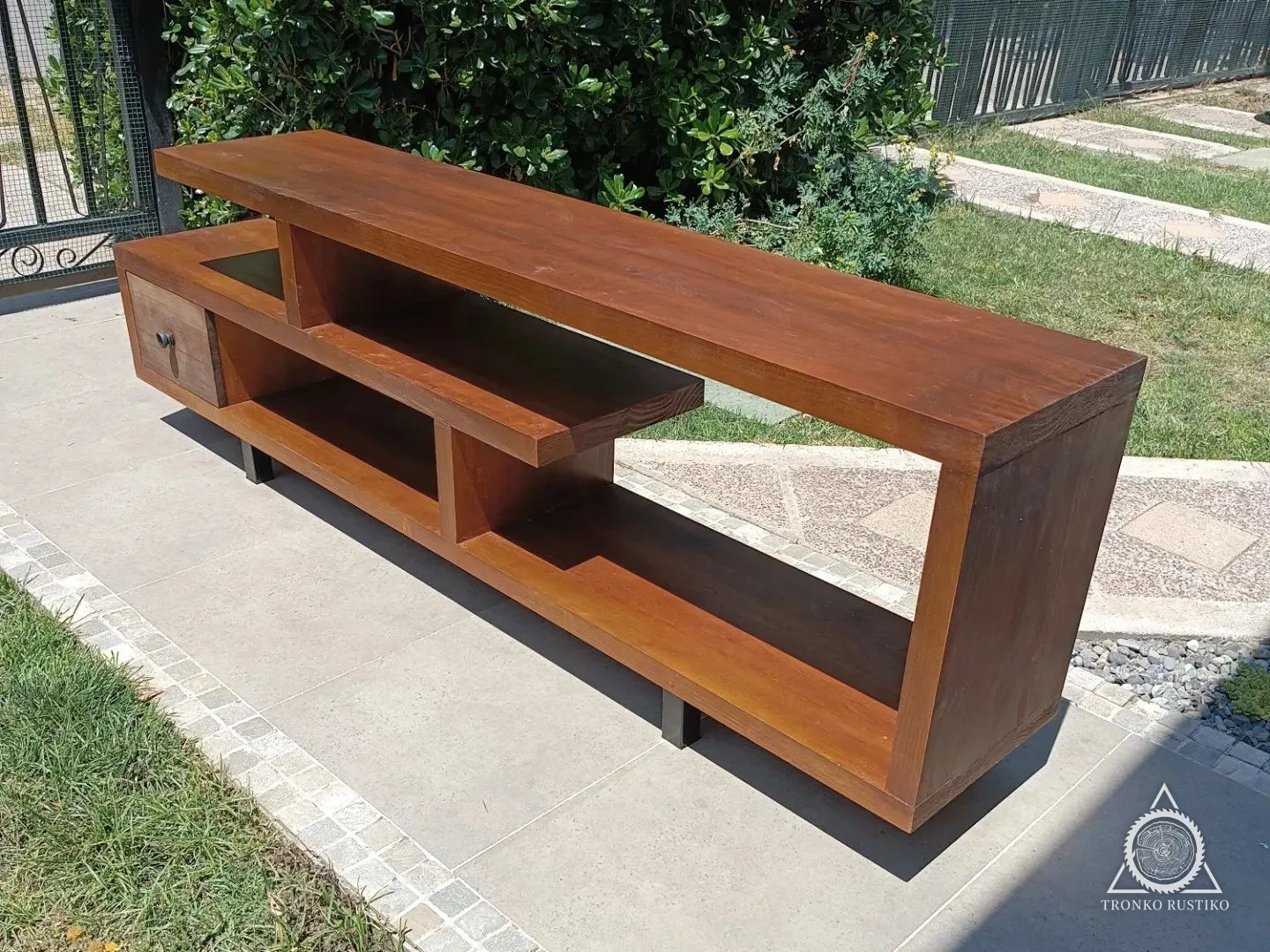 Rack bajo de madera rojiza con planos horizontales desplazados, nichos abiertos y un cajón, sobre patas negras, en un jardín.
