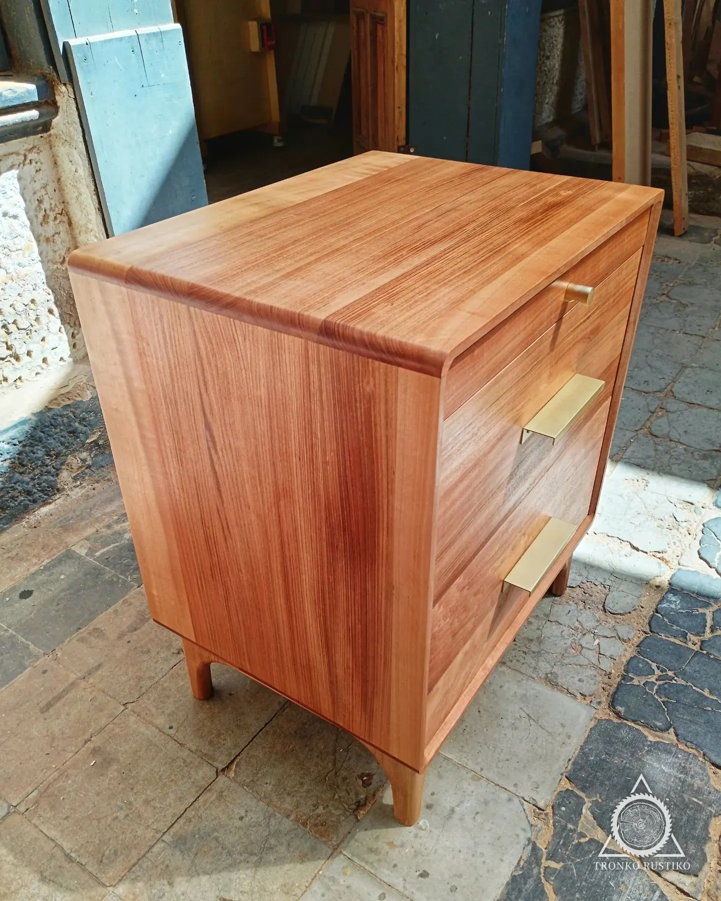 Velador de madera rojiza con cantos curvos, cajones con tiradores dorados y patas curvas.