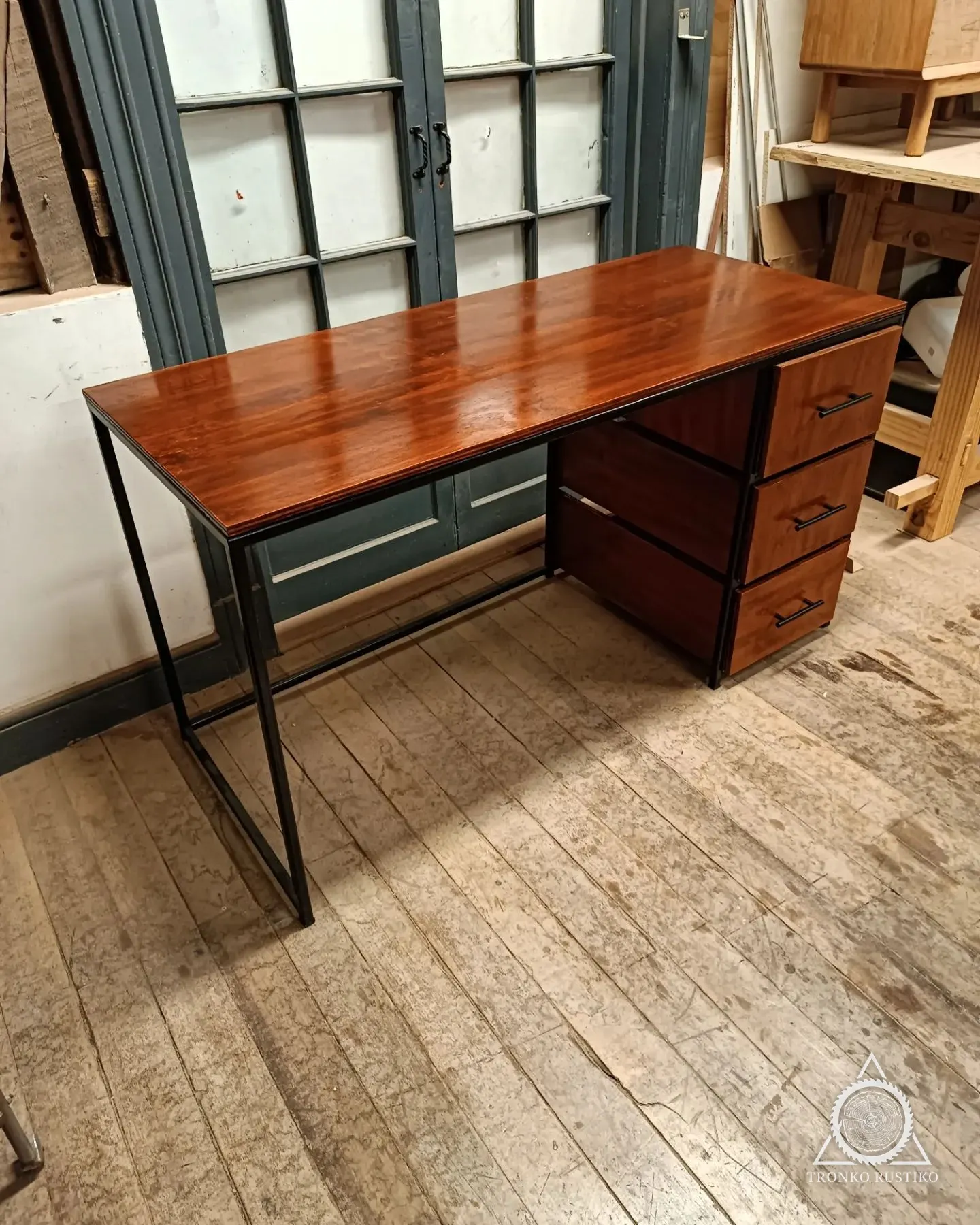 Escritorio de madera rojiza con estructura metálica negra y cajonera de tres cajones.