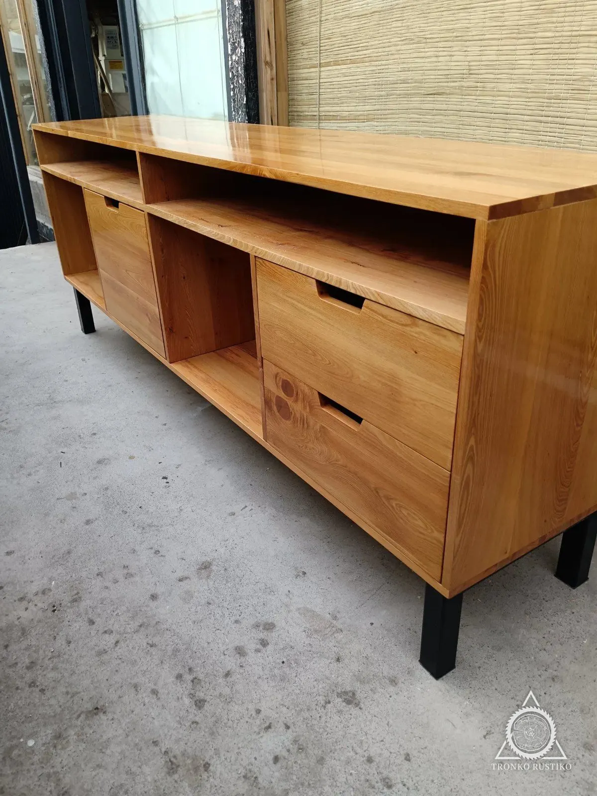 Rack de madera clara con nichos abiertos arriba, una puerta y cajones con tiradores rebajados, sobre patas metálicas negras.