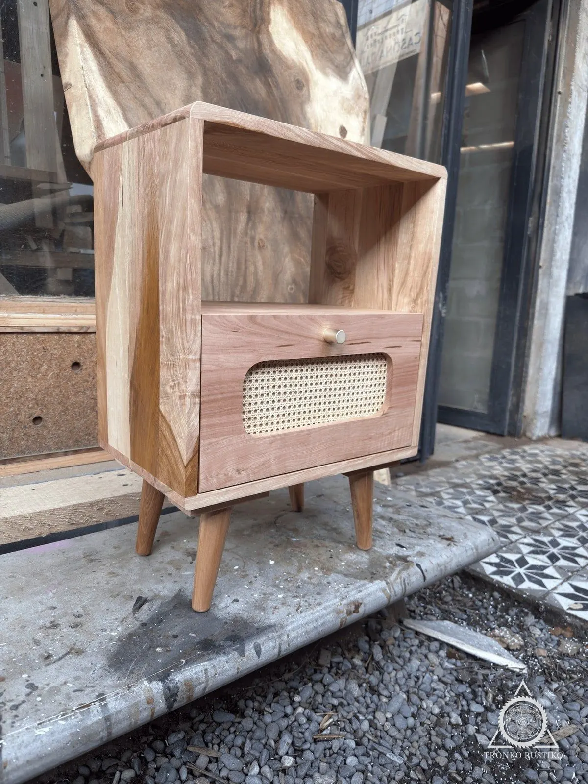 Velador de madera clara con nicho abierto y cajón de frente de ratán, sobre patas inclinadas, en la entrada del taller.