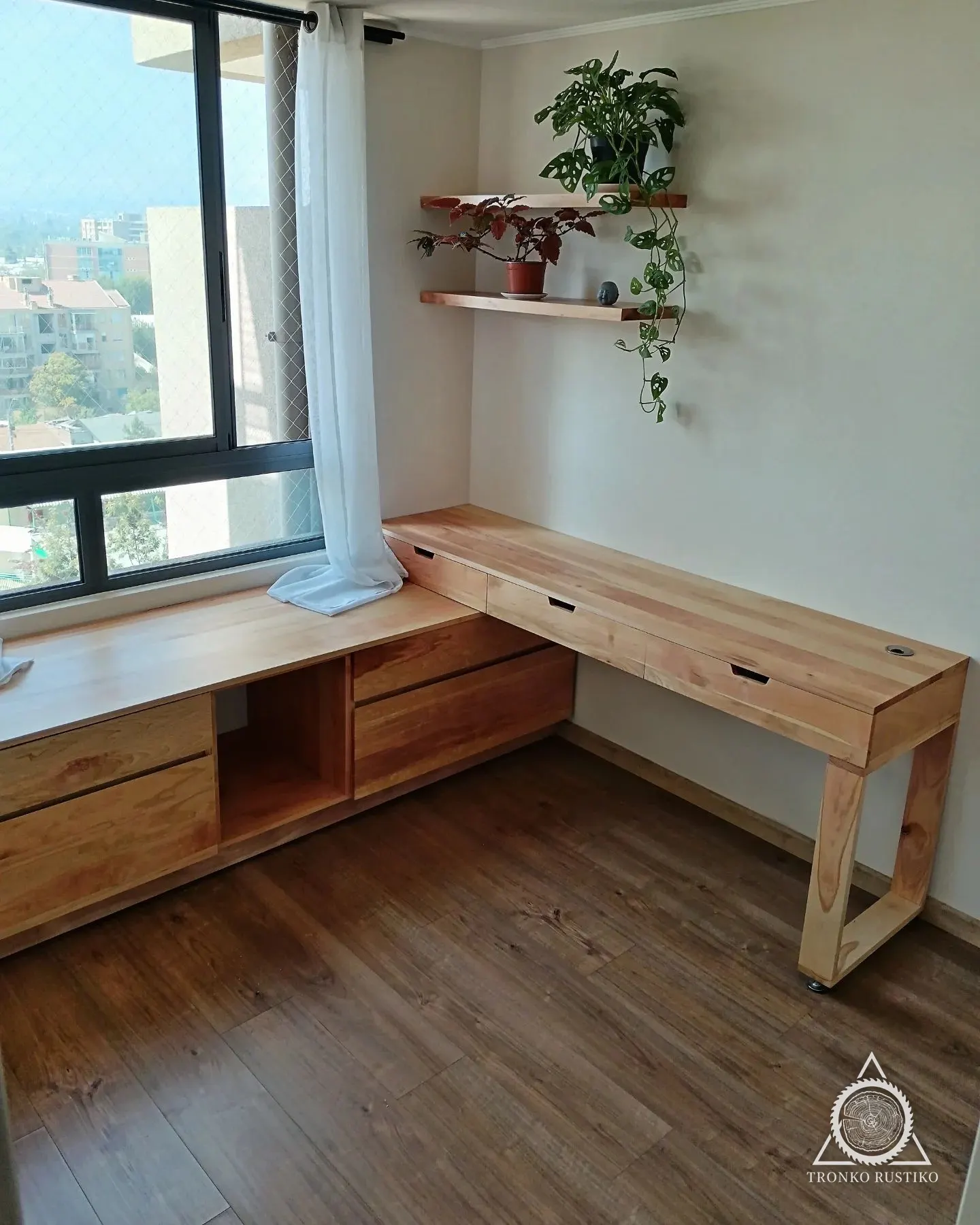 Escritorio en L de madera clara bajo una ventana, con cajonera integrada y repisas flotantes con plantas en el muro.