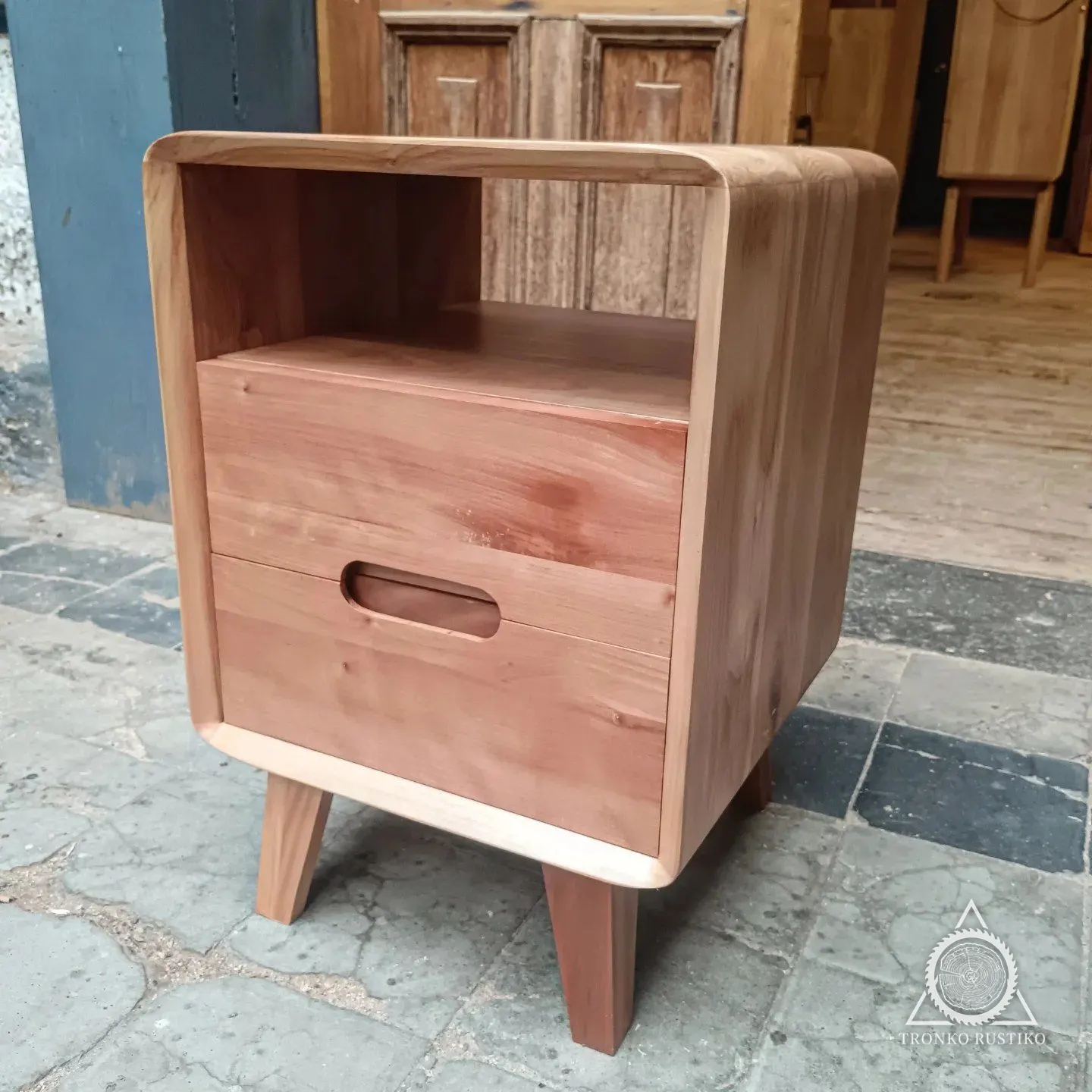 Velador de madera clara de cantos curvos con nicho abierto arriba, cajón con tirador calado y patas inclinadas.