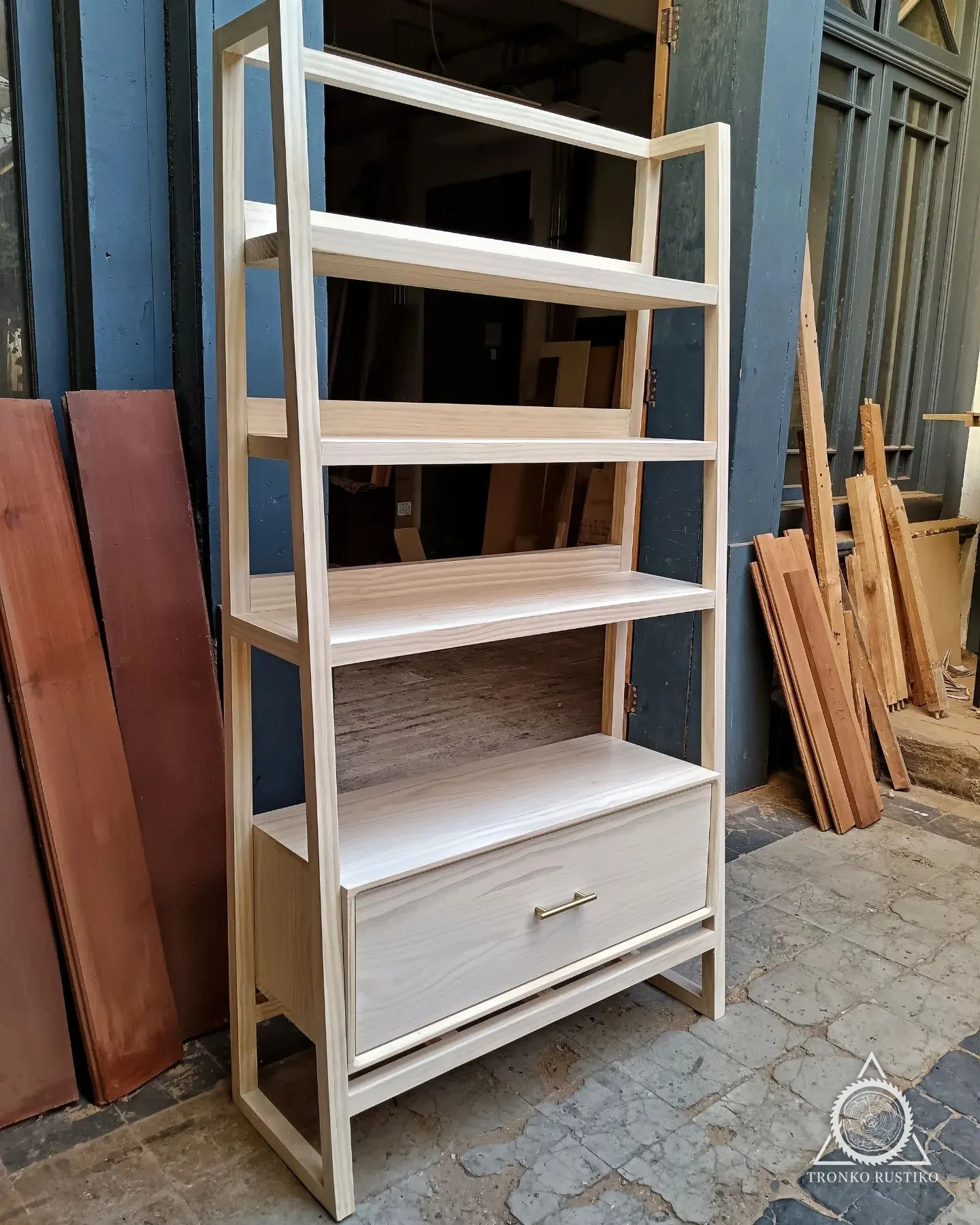 Librero de madera clara con repisas abiertas, estructura inclinada y cajón inferior.