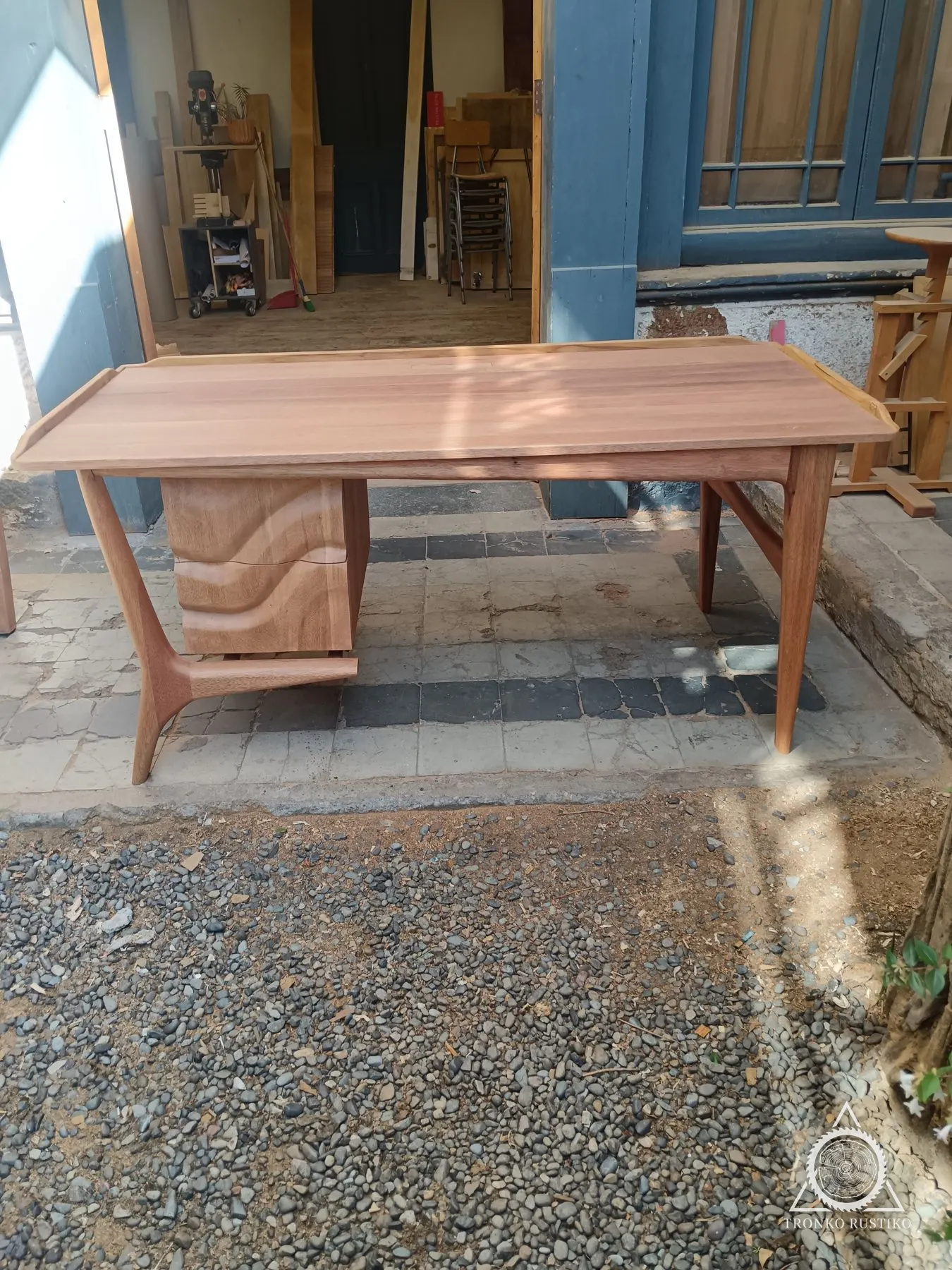Escritorio de madera de estilo mid-century con patas esculpidas y cajones suspendidos de frente tallado, en el patio del taller.
