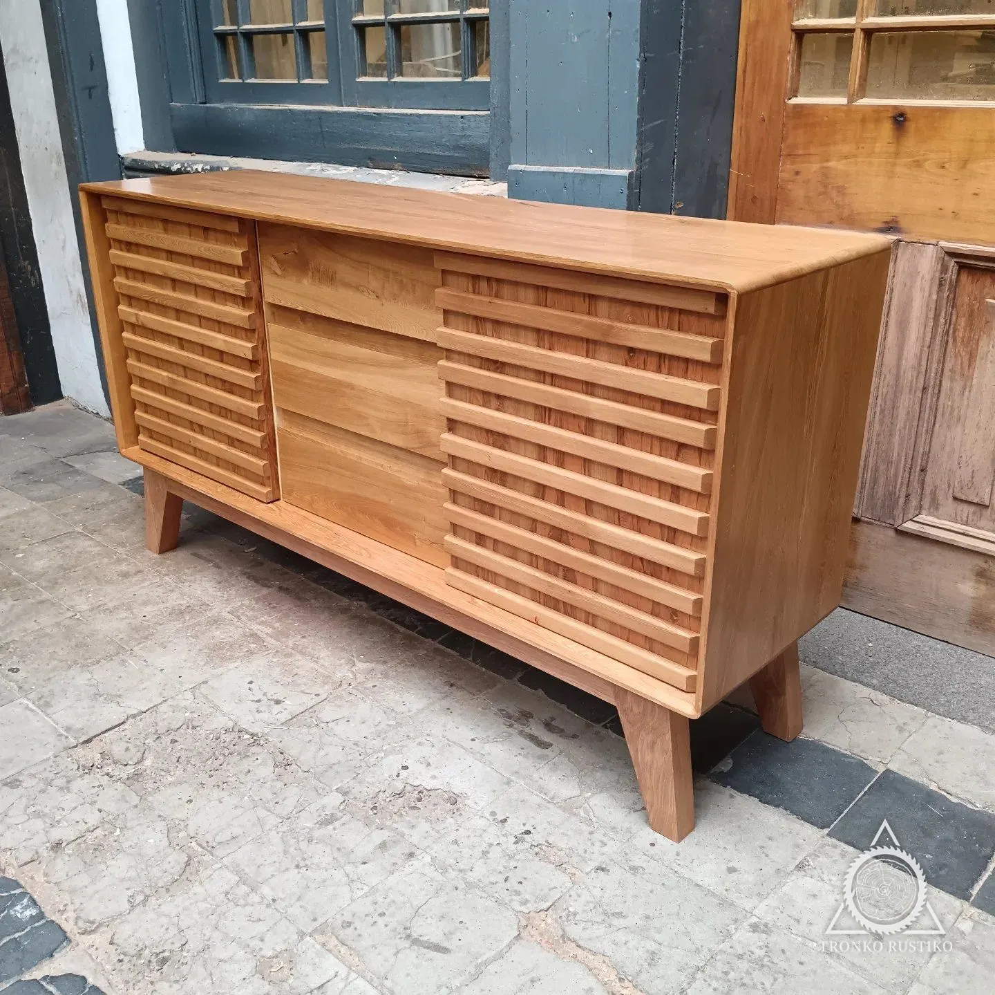 Rack de madera clara con puertas de listones horizontales a los lados, tres cajones al centro y patas inclinadas, frente al taller.