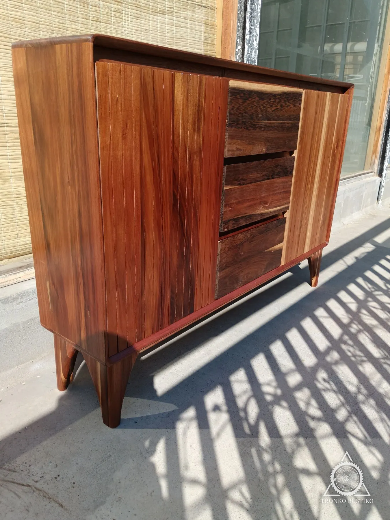 Arrimo de madera rojiza con puertas de palillaje vertical y cajones al centro, sobre patas curvas.